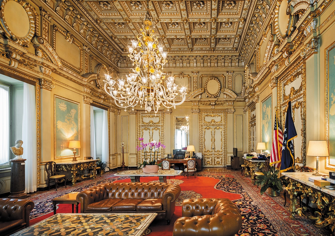 Ornate gilded reception room inside the U.S. Chancery in Rome, with a crystal chandelier and American flags
